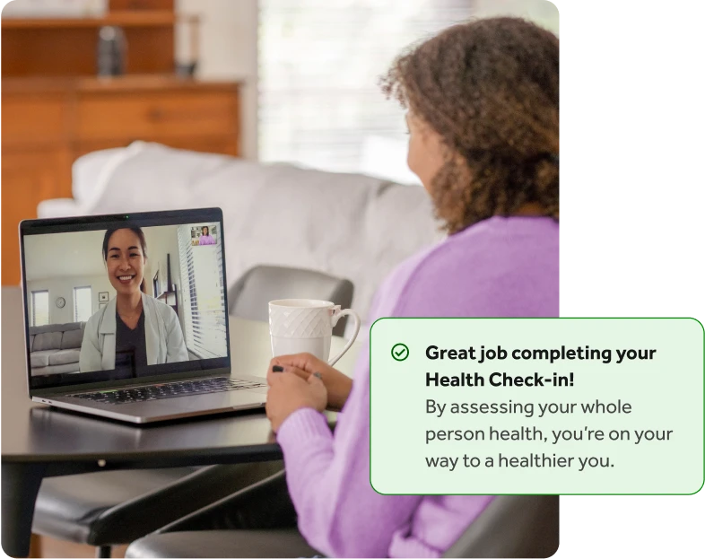 Woman with curly hair in a purple shirt talking to a doctor on her laptop at a table, with an overlay of a Teladoc Health health check-in completion notification.